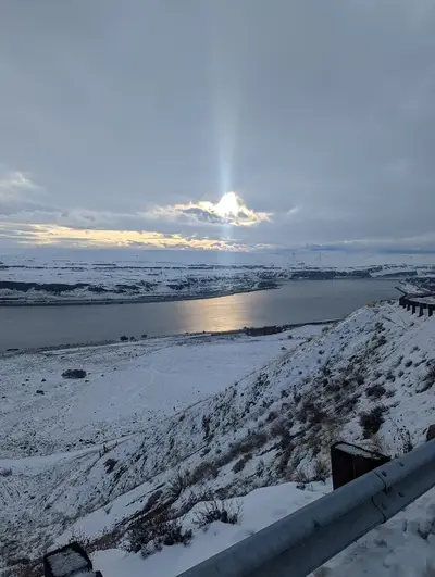 A snowy river valley with a highway and rail line running alongside the water at sunset
